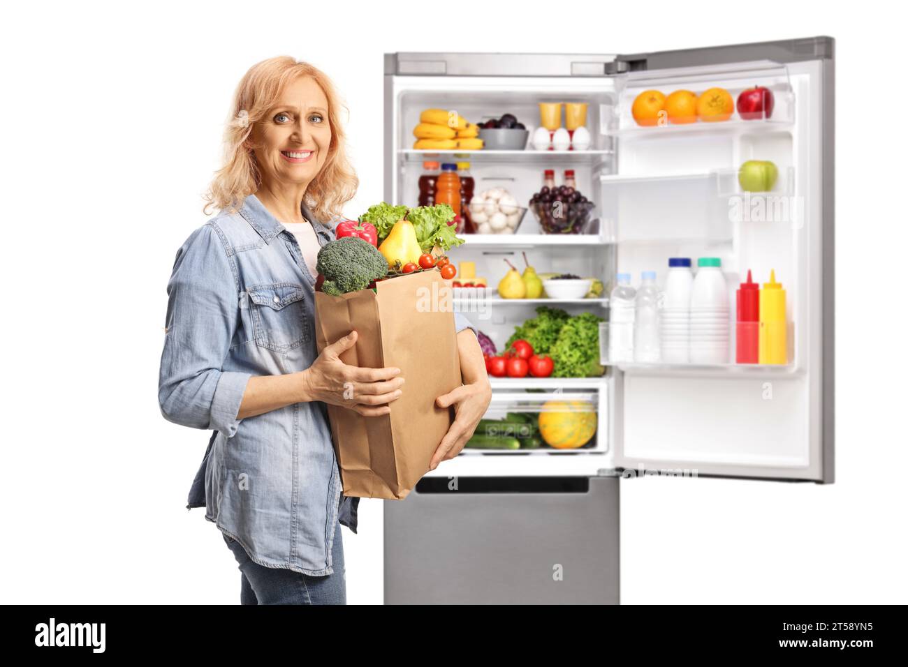Reife Frau mit einem Lebensmittelbeutel neben einem offenen Kühlschrank mit Essen isoliert auf weißem Hintergrund Stockfoto