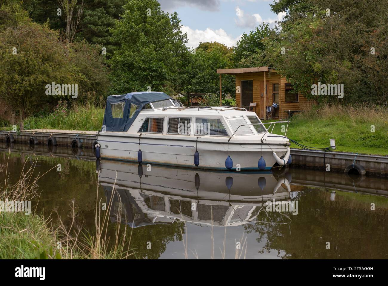 Ein schmales Boot liegt auf einem Kanal vor einem Holzschuppen. Es spiegelt sich perfekt im stillen Wasser wider Stockfoto