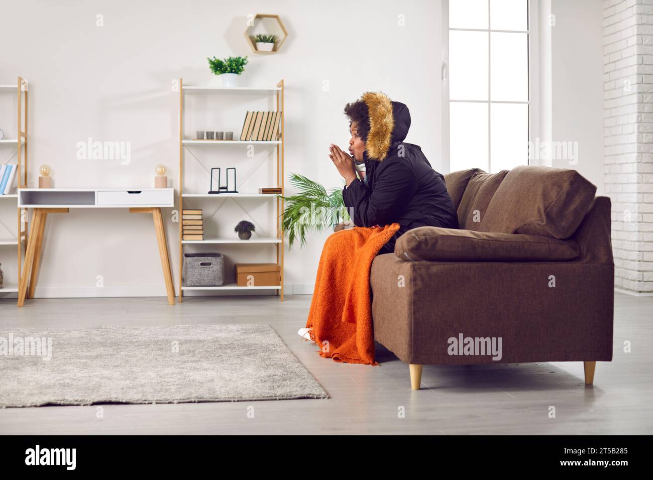 Junge gefrorene Frau, die zu Hause in einem Wintermantel auf dem Sofa sitzt. Heizungsprobleme Konzept. Stockfoto