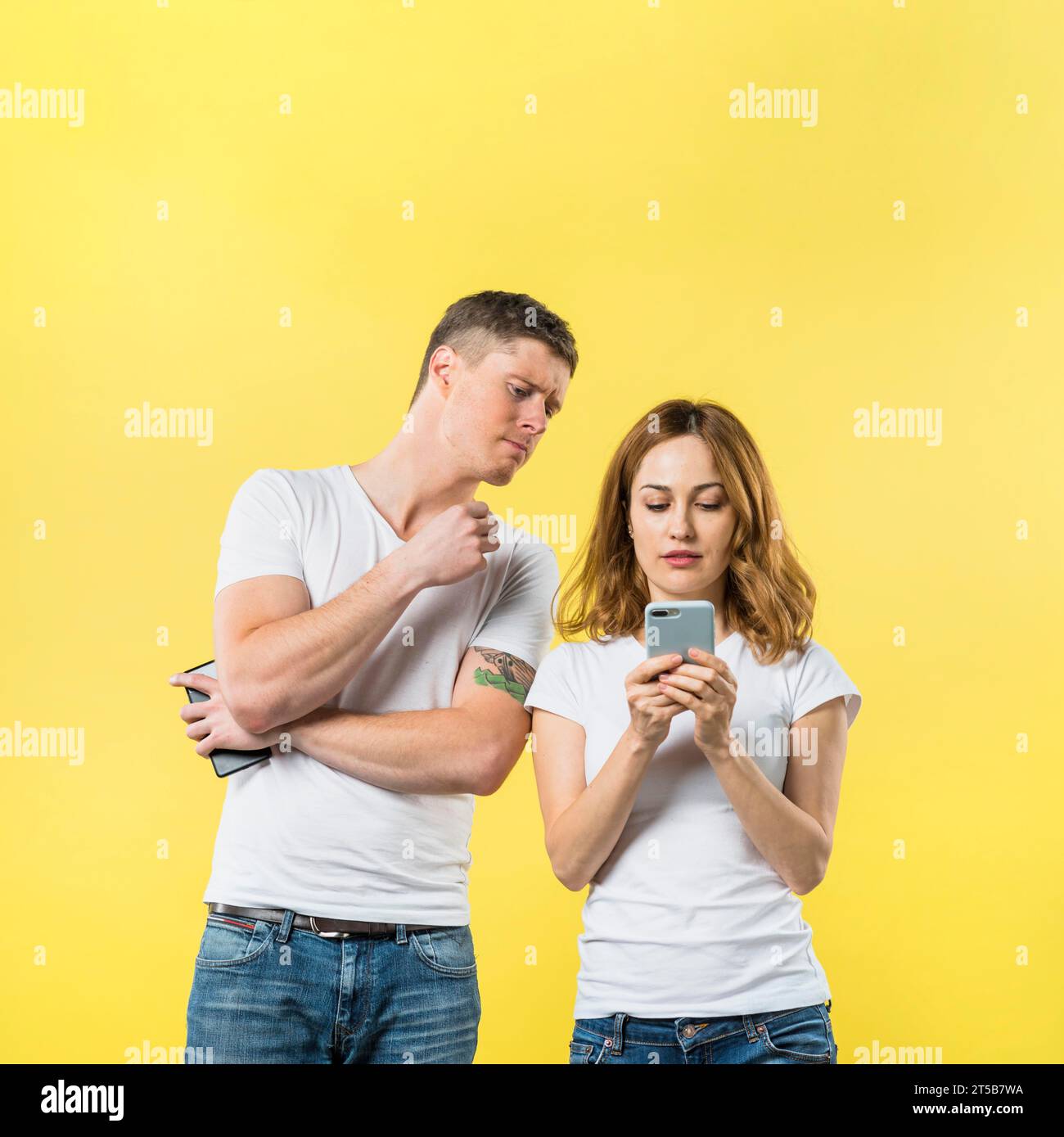 Eifersüchtiger Freund, der seine Freundin ausspioniert und ihr Handy vor gelbem Hintergrund beobachtet Stockfoto