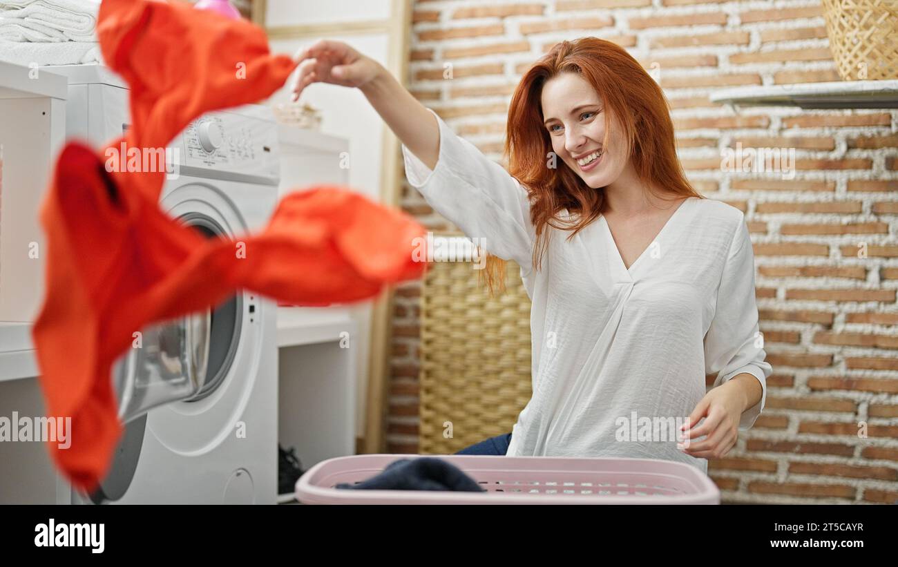 Die junge rothaarige Frau wirft Kleidung in die Luft und lächelt in der Wäscherei Stockfoto