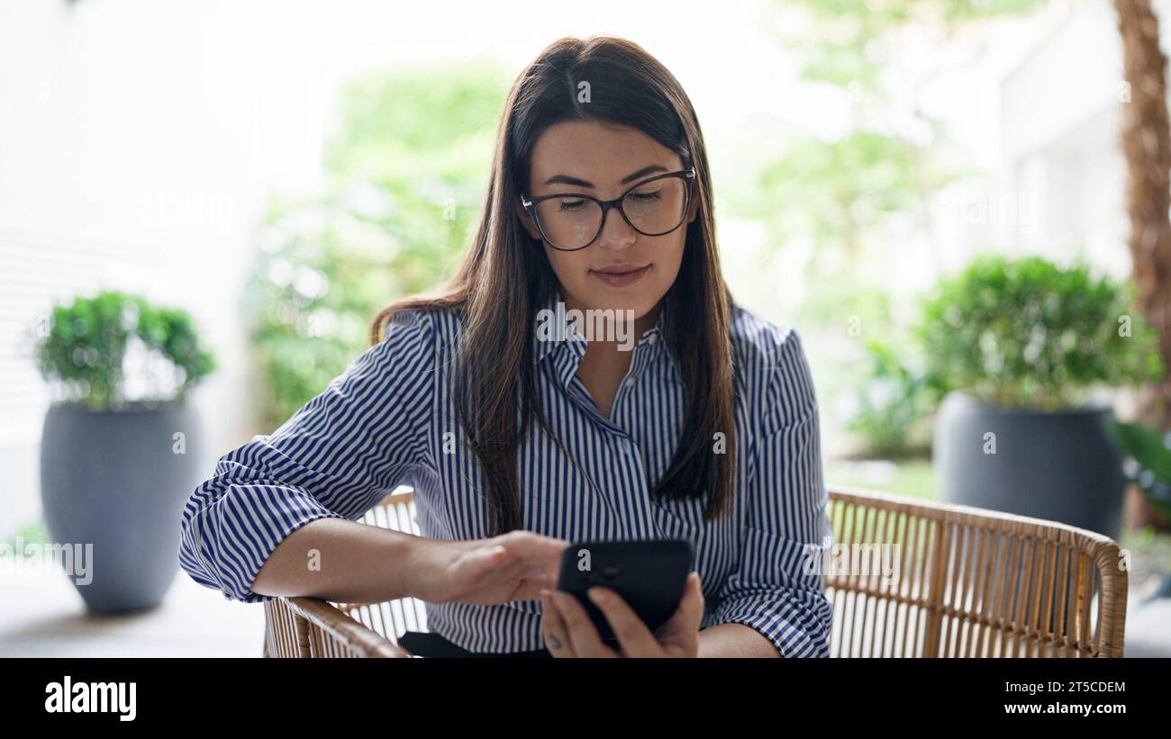 Junge, wunderschöne hispanische Frau, die mit ihrem Smartphone lächelt, sitzt auf einem Stuhl im Bürogarten Stockfoto