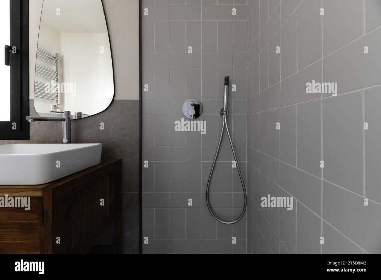 Kleines, modernes Badezimmer mit grauen Fliesen in Steinoptik, Spiegel an der Wand, weißes Porzellanwaschbecken, das über Holzschrank auskragt, Duschkabine mit gla Stockfoto