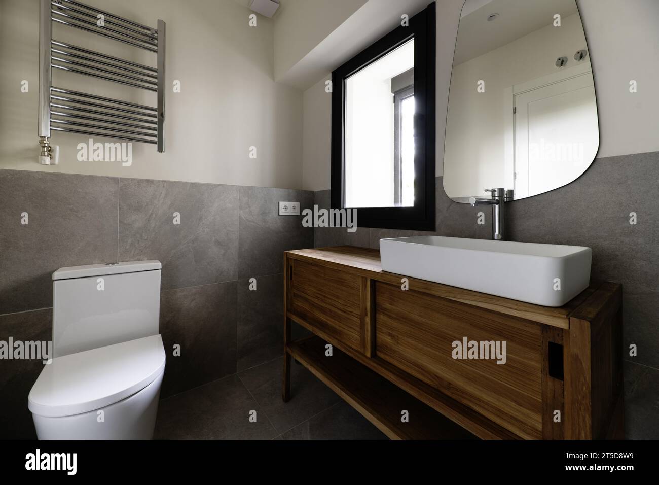 Kleines, modernes Design-Badezimmer mit grauen Fliesen mit Steineffekt, Spiegel an der Wand, weißes Porzellanwaschbecken, das auf einem Holzschrank auskragt, Handtuchtrocknung radia Stockfoto