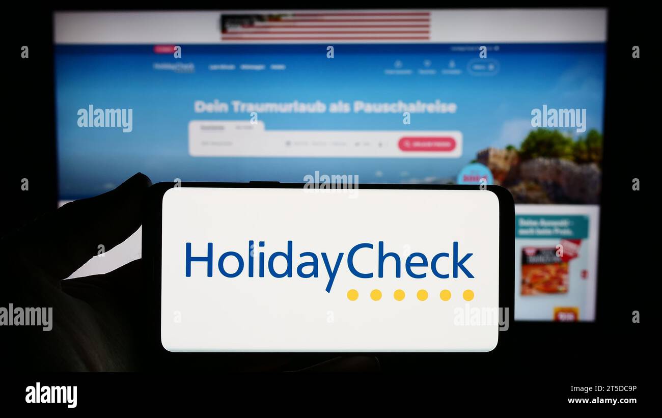 Person mit Smartphone mit Logo der Schweizer Reisebuchungsportalgesellschaft HolidayCheck AG vor der Website. Schwerpunkt auf dem Display des Telefons. Stockfoto