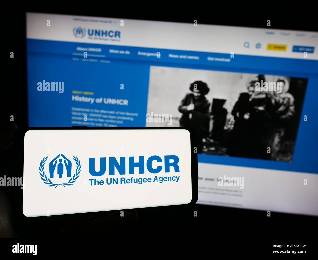 Person, die ein Mobiltelefon mit dem Logo des Hohen Flüchtlingskommissars der Vereinten Nationen (UNHCR) vor der Webseite hält. Schwerpunkt auf dem Display des Telefons. Stockfoto