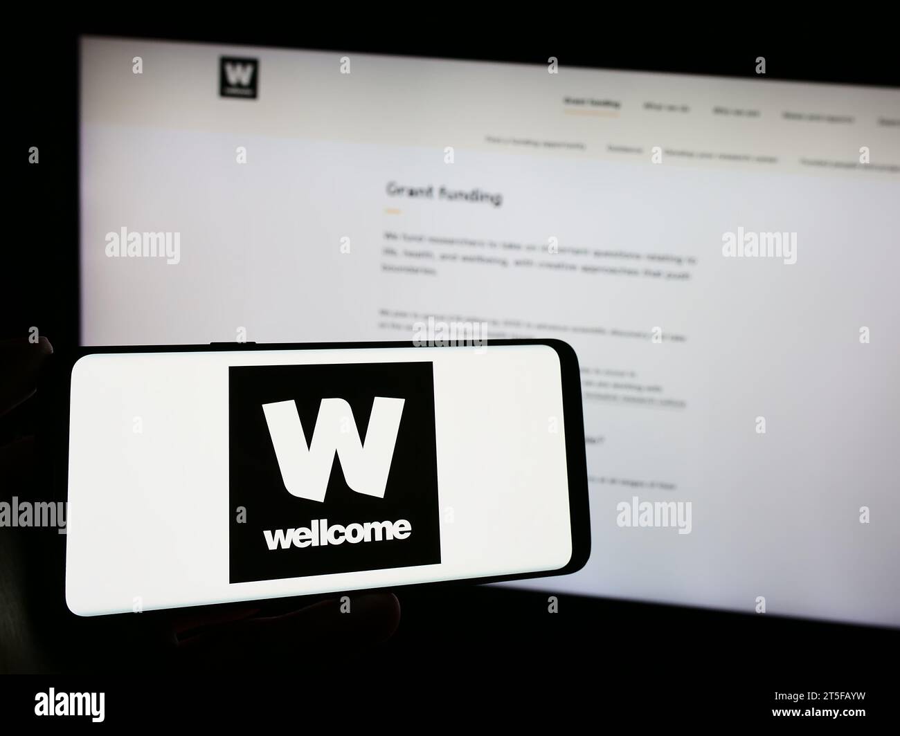 Person, die ein Mobiltelefon mit dem Logo der britischen Wohltätigkeitsstiftung Wellcome Trust vor der Webseite hält. Schwerpunkt auf dem Display des Telefons. Stockfoto
