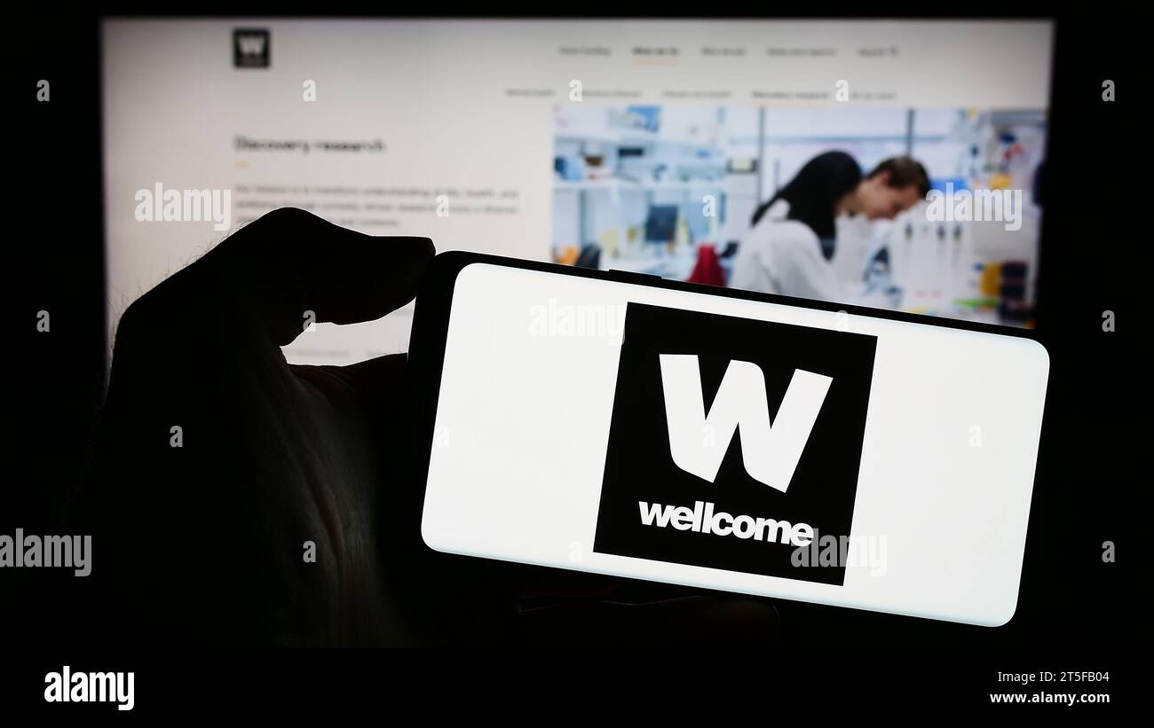 Person mit Handy mit Logo der britischen Wohltätigkeitsstiftung Wellcome Trust vor der Webseite. Schwerpunkt auf dem Display des Telefons. Stockfoto