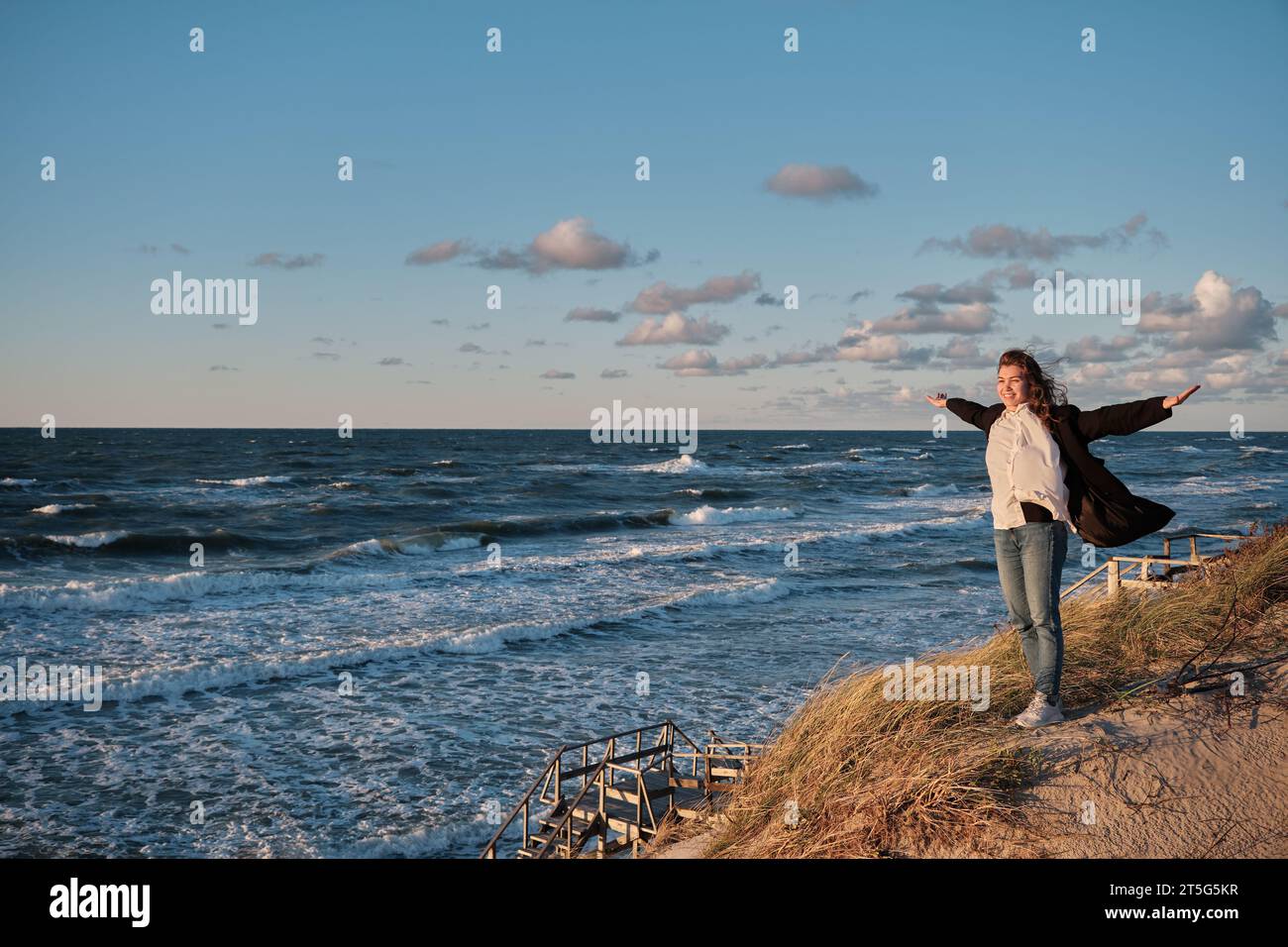 Glückliche Frau steht an der Sandküste in der Nähe des stürmischen Meeres und blickt in die Kamera Stockfoto