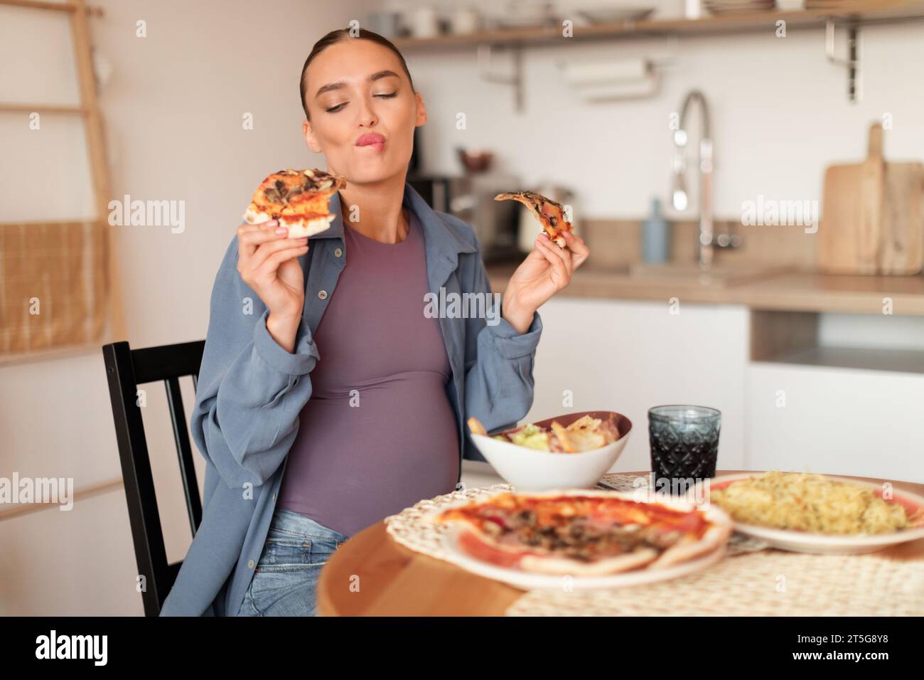 Schwangere Frau, die zwei Pizzascheiben in der Küche schätzt Stockfoto