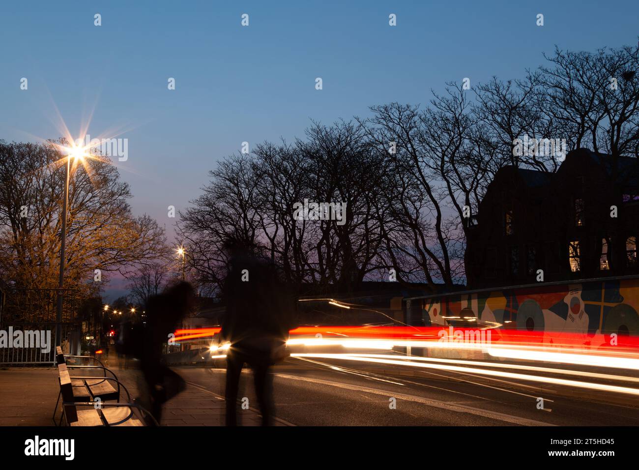 Eine Langzeitaufnahme von Lichtspuren, die von Autolichtern mit Silhouetten von Menschen auf einer Bank auf der Mill Road Bridge in Cambridge, England, Großbritannien, erzeugt wurden. Stockfoto