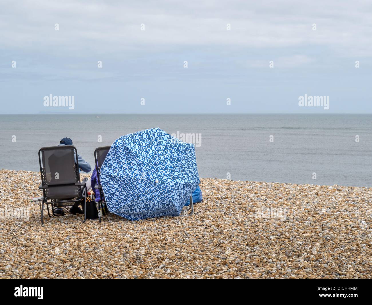 LYME REGIS, DORSET, Großbritannien - 7. MAI 2023: Der Inbegriff eines englischen Sommers, in dem sich ein Paar in Winterkleidung unter einem Sonnenschirm versteckt. Stockfoto