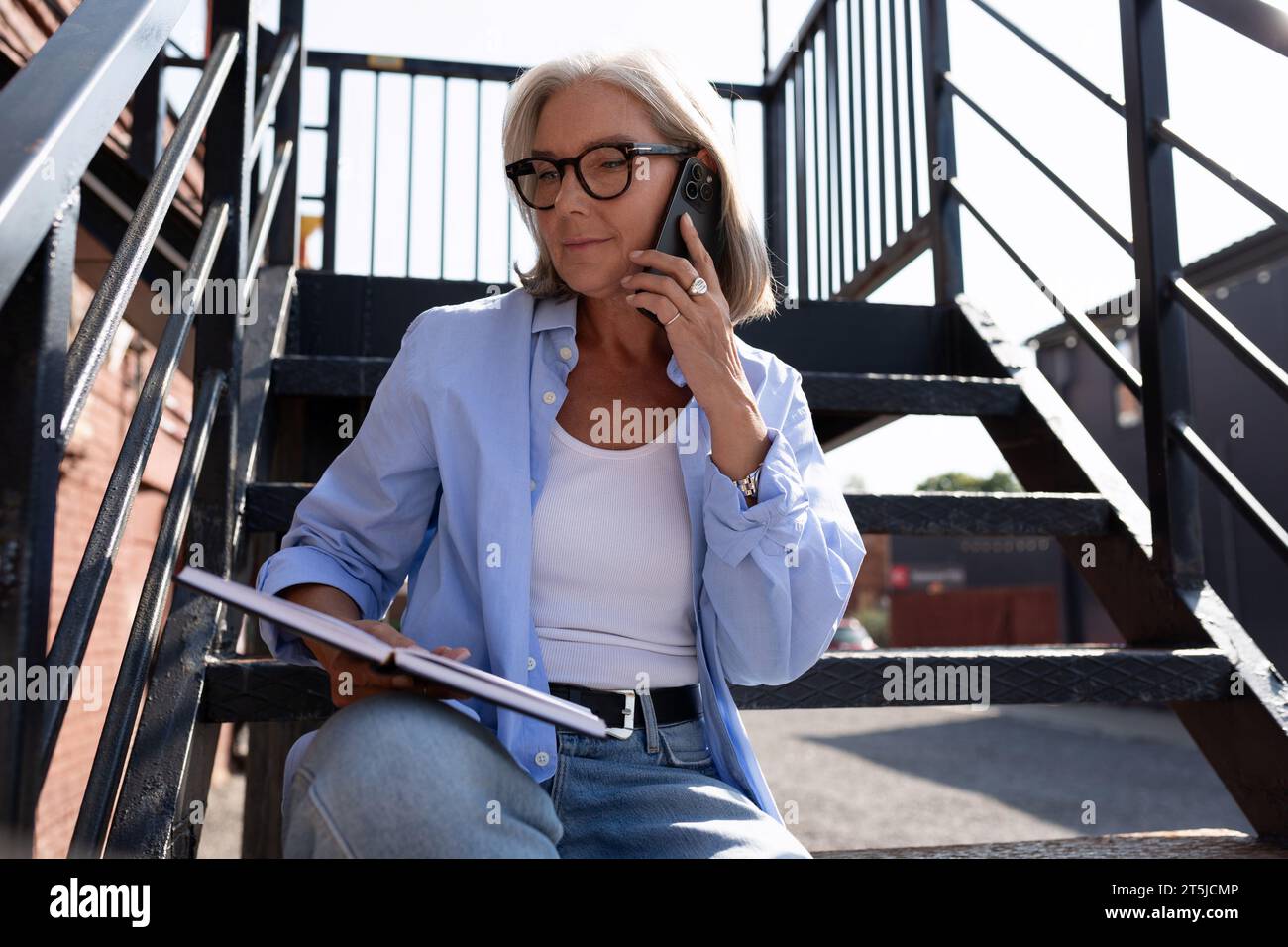 Reife grauhaarige Frau im Sommeroutfit arbeitet mit einem Laptop, während sie in der Stadt auf der Straße sitzt Stockfoto