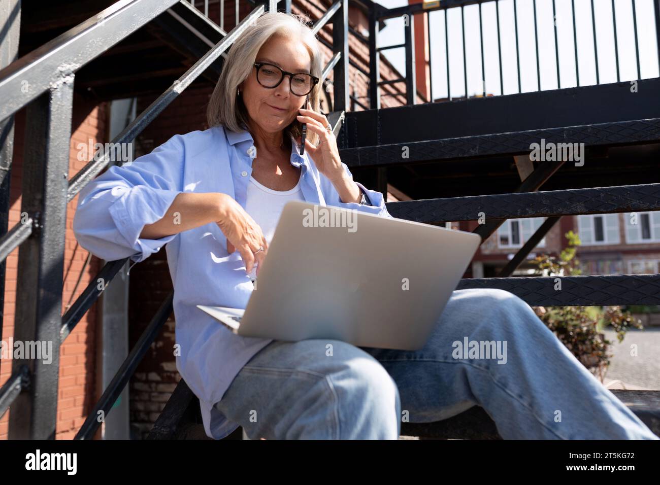 Reife grauhaarige Frau im Sommeroutfit arbeitet mit einem Laptop, während sie in der Stadt auf der Straße sitzt Stockfoto