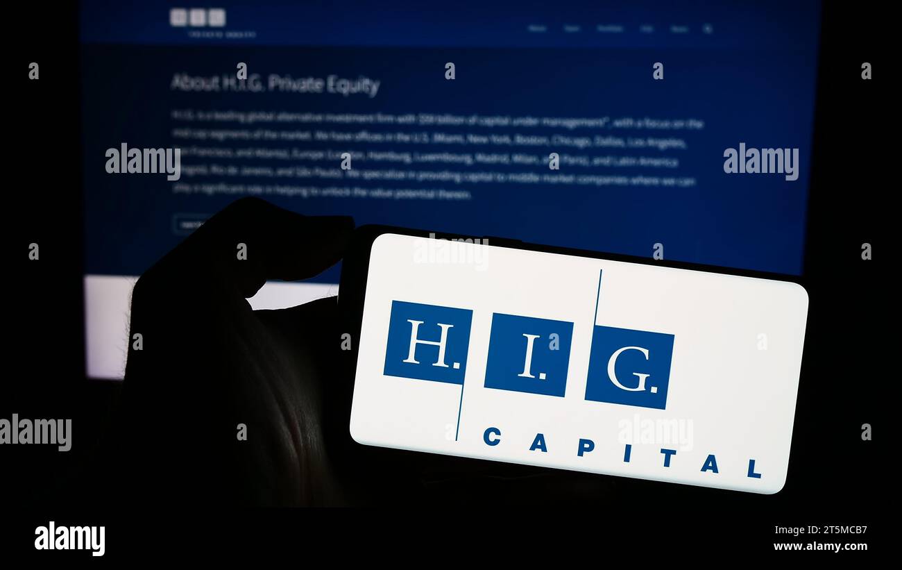 Person, die ein Mobiltelefon mit dem Logo der amerikanischen Investmentgesellschaft H.I.G. Capital LLC vor der Webseite hält. Schwerpunkt auf dem Display des Telefons. Stockfoto