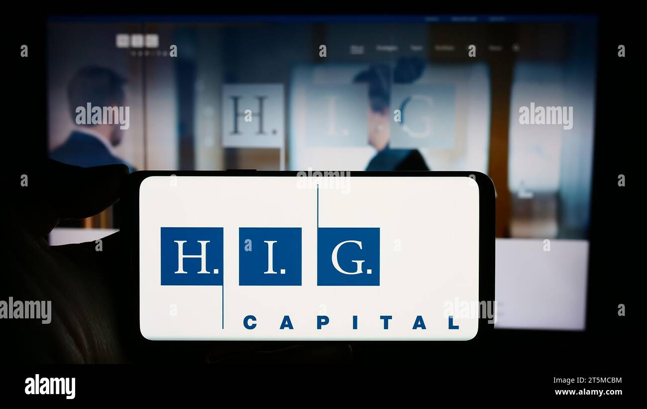 Person, die ein Mobiltelefon mit dem Logo der US-Investmentgesellschaft H.I.G. Capital LLC vor der Website des Unternehmens hält. Schwerpunkt auf dem Display des Telefons. Stockfoto