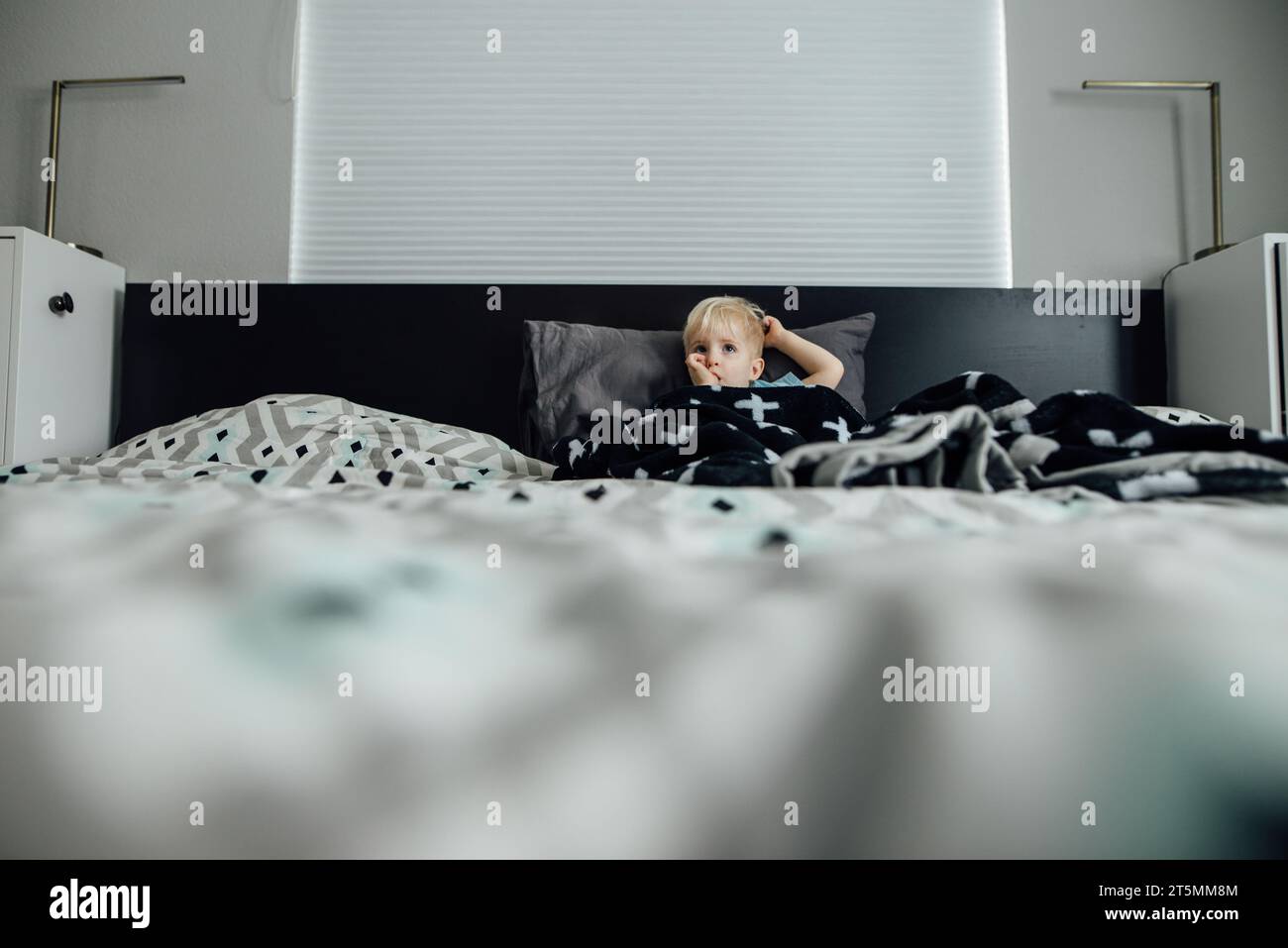 Großer Blick auf einen kleinen Jungen, der auf einem großen Bett liegt und seinen Daumen lutscht Stockfoto