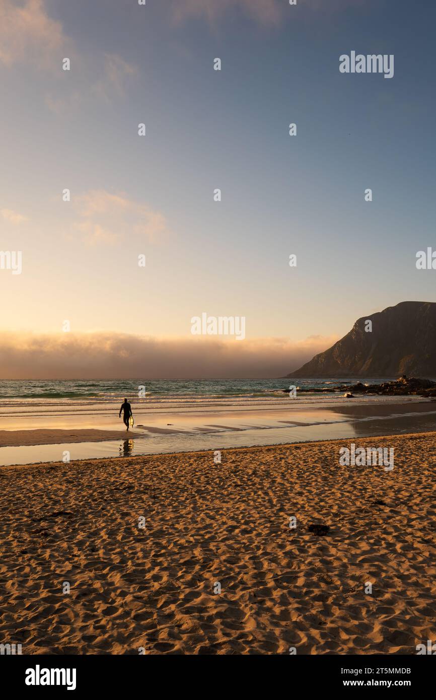 Surfer bei Sonnenuntergang an einem Strand in Lofoten, Norwegen, unter der Mitternachtssonne. Blauer Himmel mit Nebel, der hereinrollt. Stockfoto