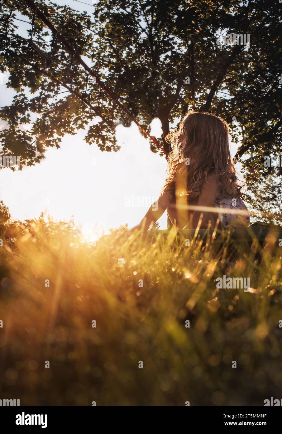 Kleines Mädchen mit langen Locken, das bei Sonnenuntergang im Gras sitzt Stockfoto