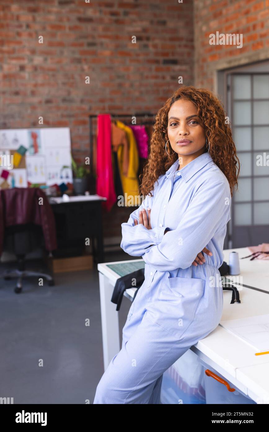 Glückliche, birassische Modedesignerin, die sich mit gefalteten Armen auf dem Schreibtisch in einem sonnigen Studio stützt Stockfoto