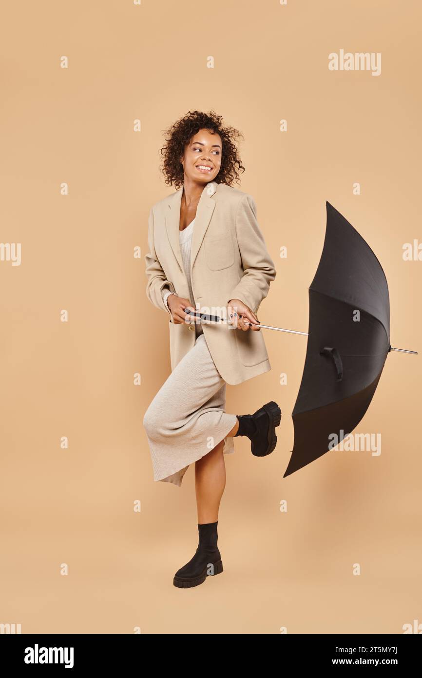 Fröhliche afroamerikanische Frau in Midikleid und herbstlichem Blazer mit Regenschirm auf Beige Stockfoto