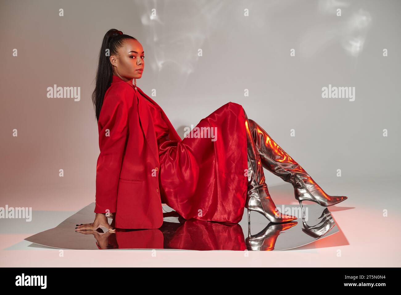 Junge afroamerikanische Frau in rotem Kleid, Blazer und silbernen Stiefeln, die auf einer spiegelnden Oberfläche sitzt Stockfoto