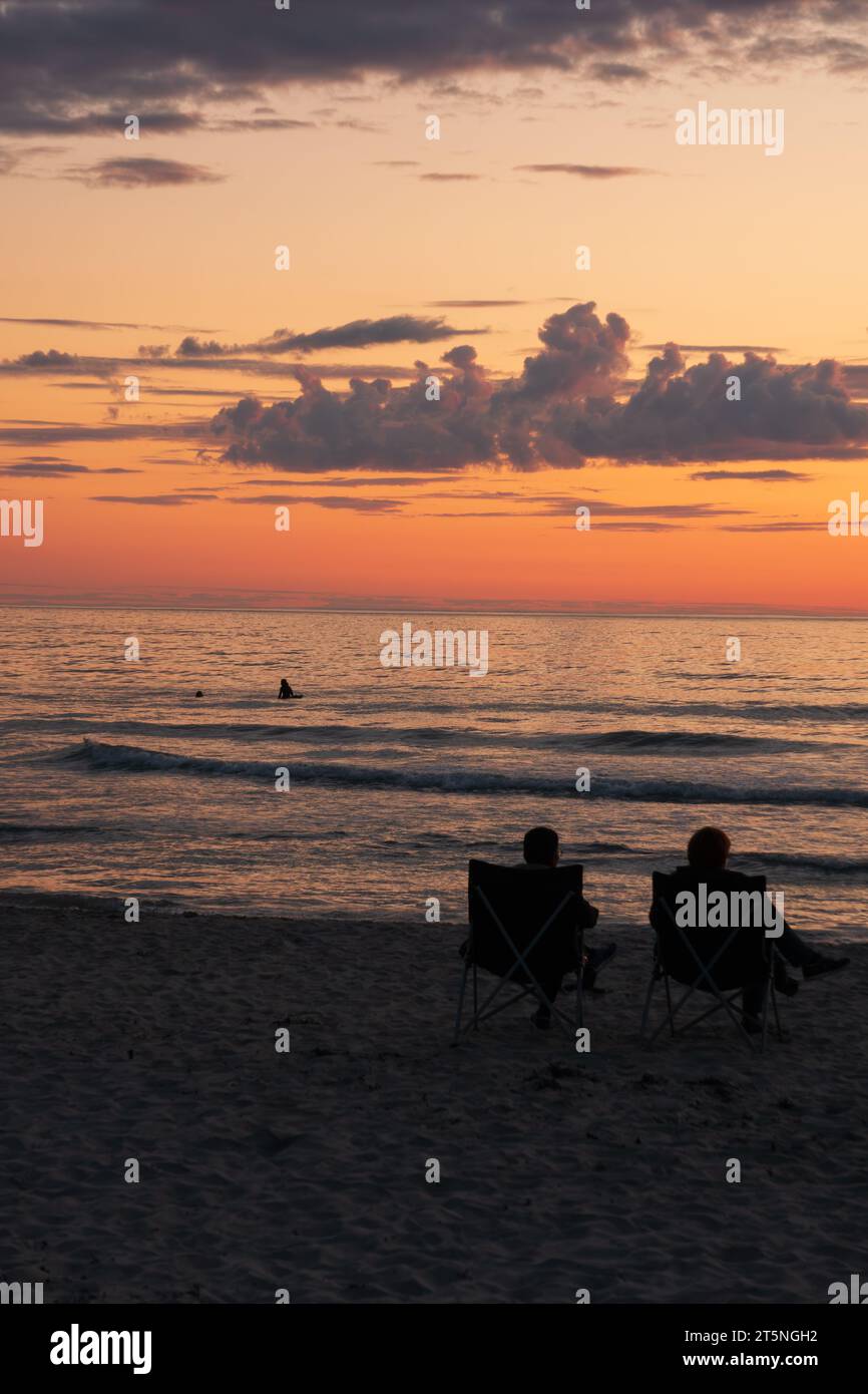 Zwei Personen sitzen in Stühlen und beobachten die Mitternachtssonne über dem Meer in den nördlichen Lofoten, Norwegen, im Juli. Ein Surfer auf dem Meer Stockfoto