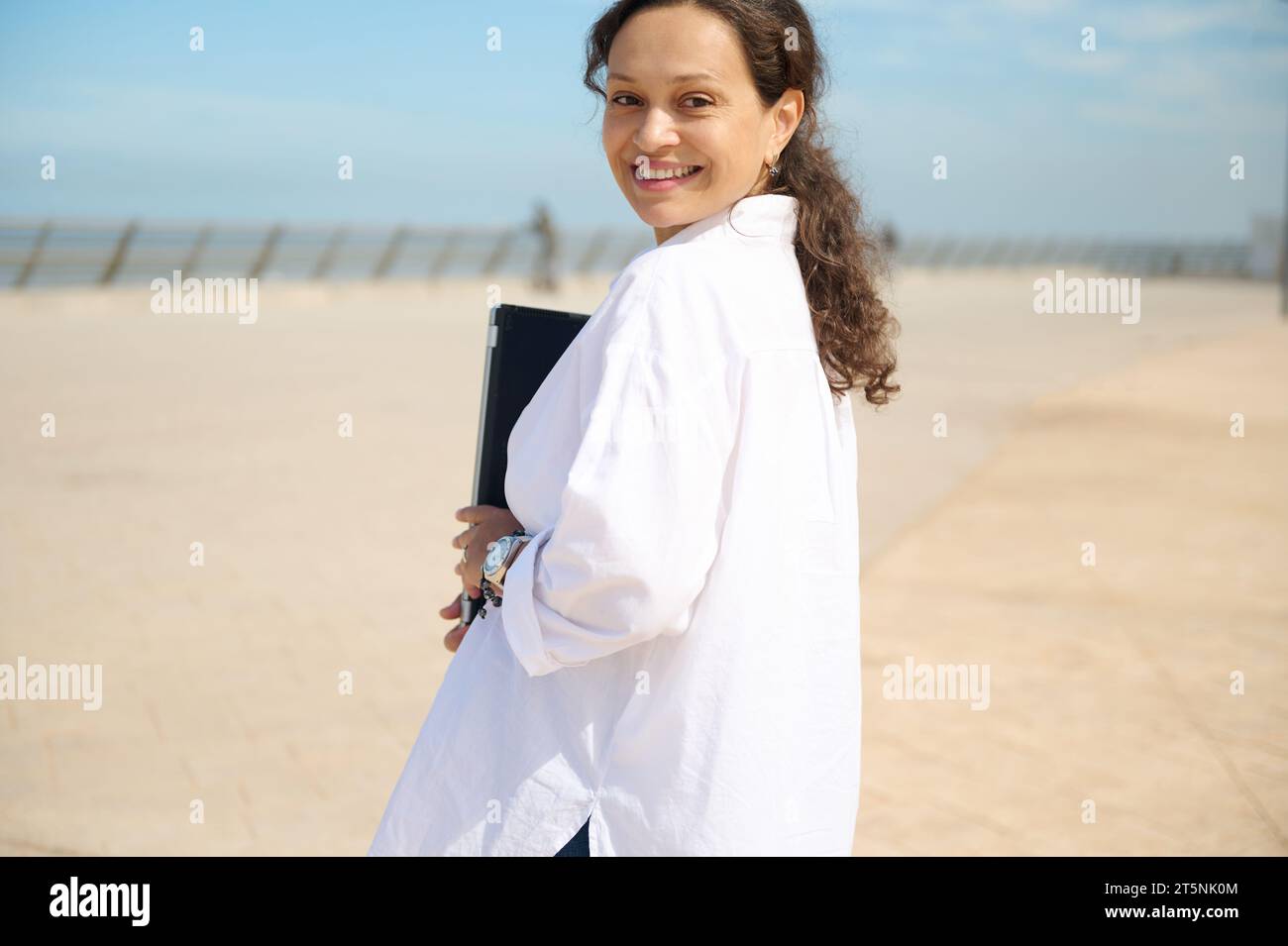 Erfolgreiche, selbstbewusste und schöne junge Multi-ethnische Geschäftsfrau in weißem Hemd, hält Laptop und blickt durch ihren Schuh in die Kamera Stockfoto