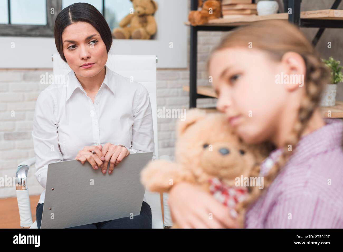 Eine Psychologin mit Klemmbrett beobachtet ein depressives Mädchen, das ihren Teddybären umarmt Stockfoto