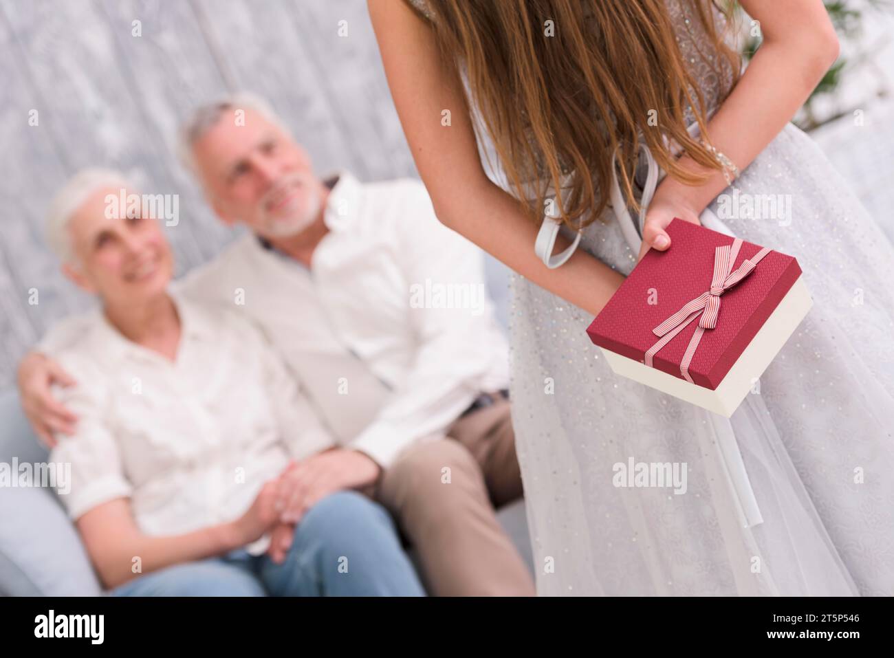 Kleines Mädchen, das ihr Geschenk versteckt, ihre Großeltern sitzen auf dem Sofa Stockfoto