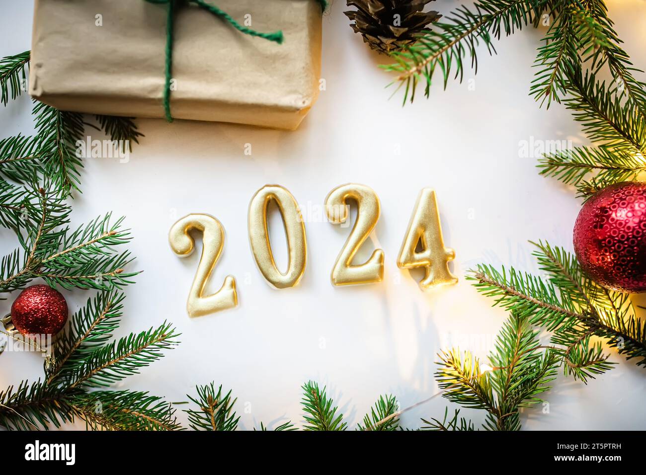 Goldene Zahlen 2024 auf weißem Hintergrund mit Bastelgeschenk, weihnachtsbaumzweigen und roten Dekorationen und weihnachtslichtern. Stockfoto