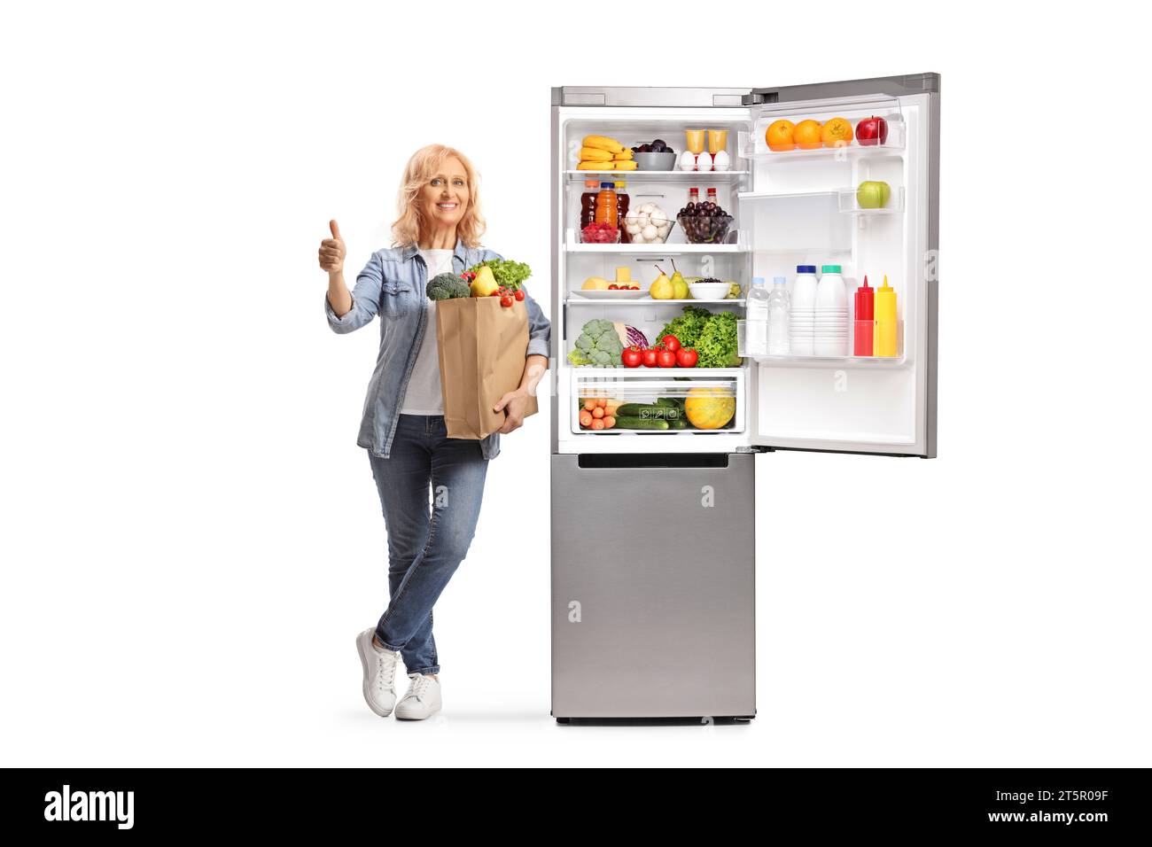 Reife Frau mit einer Einkaufstasche, die sich auf einen offenen Kühlschrank stützt und die Daumen nach oben bewegt, isoliert auf weißem Hintergrund Stockfoto
