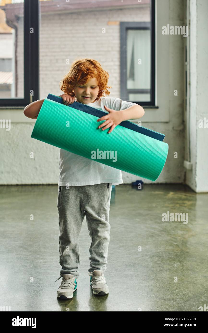 Vertikale Aufnahme eines süßen Jungen mit roten Haaren in Sportkleidung, der sein Karemat entfaltet, Kindersport Stockfoto