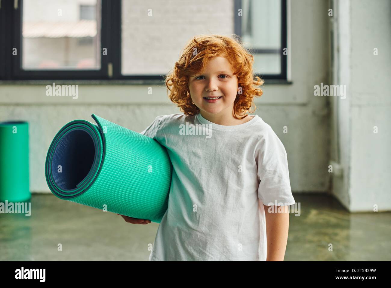 Niedlicher vorpubertärer Junge mit rotem Haar posiert mit Karemat in den Händen und lächelt in die Kamera, Kindersport Stockfoto