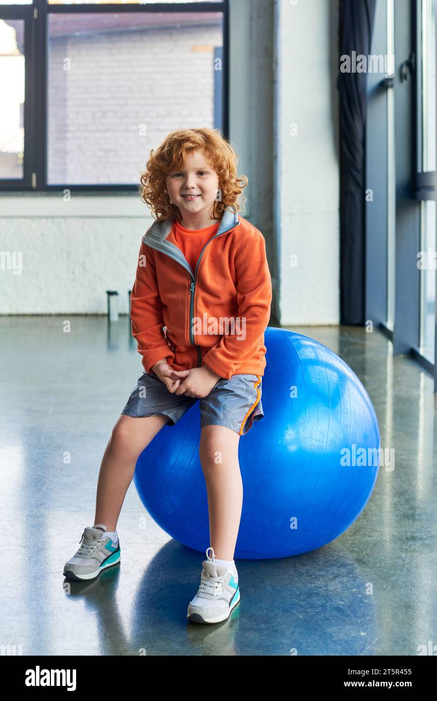 Vertikale Aufnahme eines rothaarigen, fröhlichen Jungen, der auf einem Fitnessball sitzt und freudig in die Kamera lächelt Stockfoto