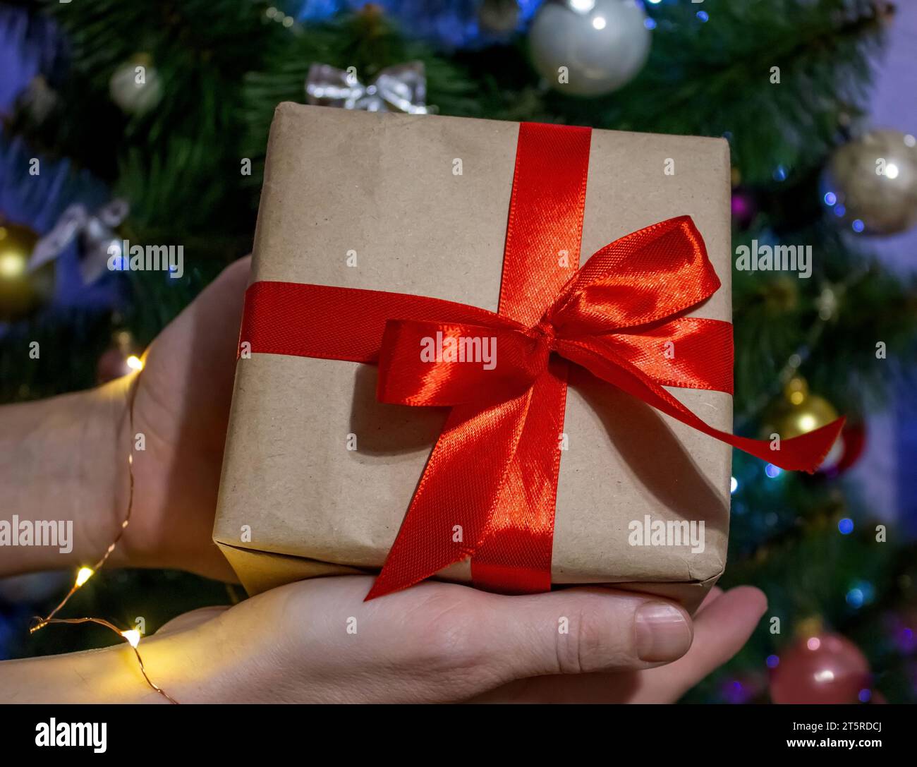Mann, der in Händen schöne Weihnachtsgeschenkbox in Kraftpapier und rotem Band auf dem Hintergrund von Weihnachtsbaum und Lichtern hält. Präsentiert Vorbereitung auf Stockfoto