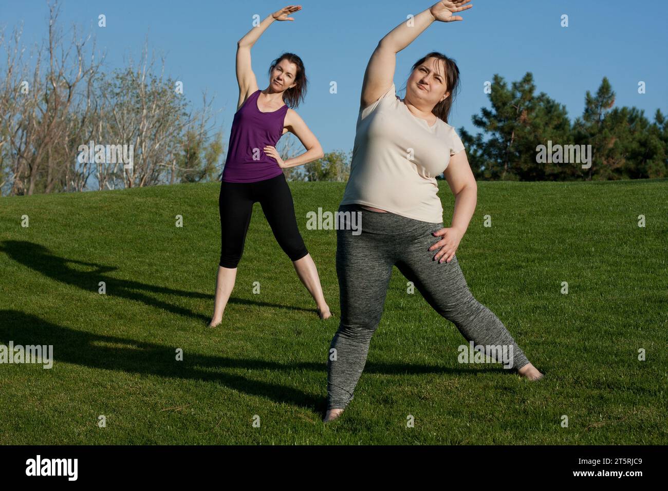 Zwei Frauen mittleren Alters trainieren im Stadtpark und beugen sich von Körper zu Seite. Gesunder Lebensstil, Yoga, Pilates, Gewichtsverlust. Übergewichtige Frau macht Yoga Witz Stockfoto