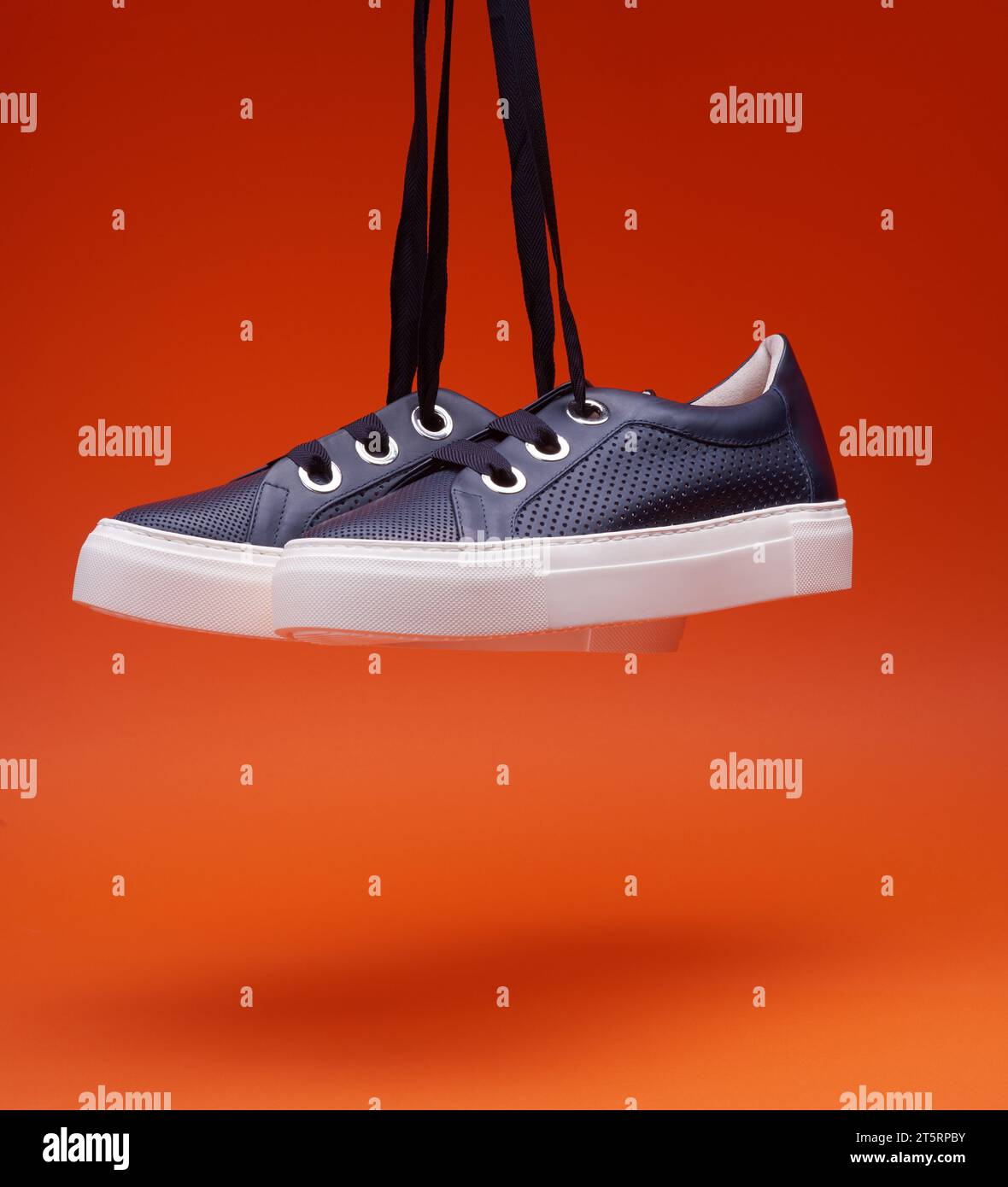 Stilvolle, legere, marineblaue Sneakers, die vor einem leuchtend orangefarbenen Hintergrund hängen. Perforiertes Leder, dicke weiße Sohle, breite Schnürsenkel. Freizeitschuhe, Stockfoto