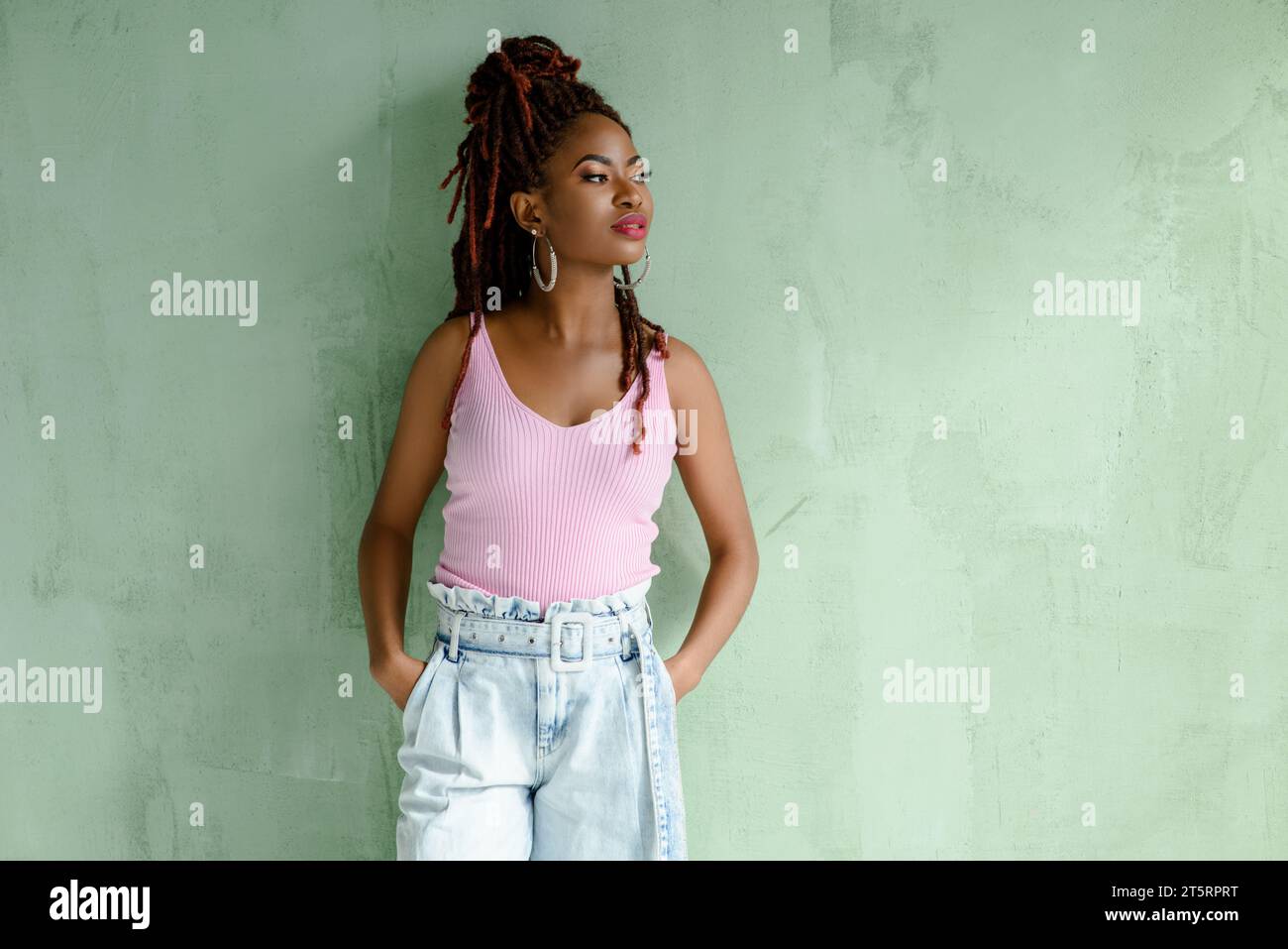 Porträt eines wunderbaren afrikanischen Mädchens mit Dreadlocks in Jeans mit weitem Bein und rosa Oberteil, wegblickend, isoliert auf hellgrüner Wand mit Kopie Stockfoto