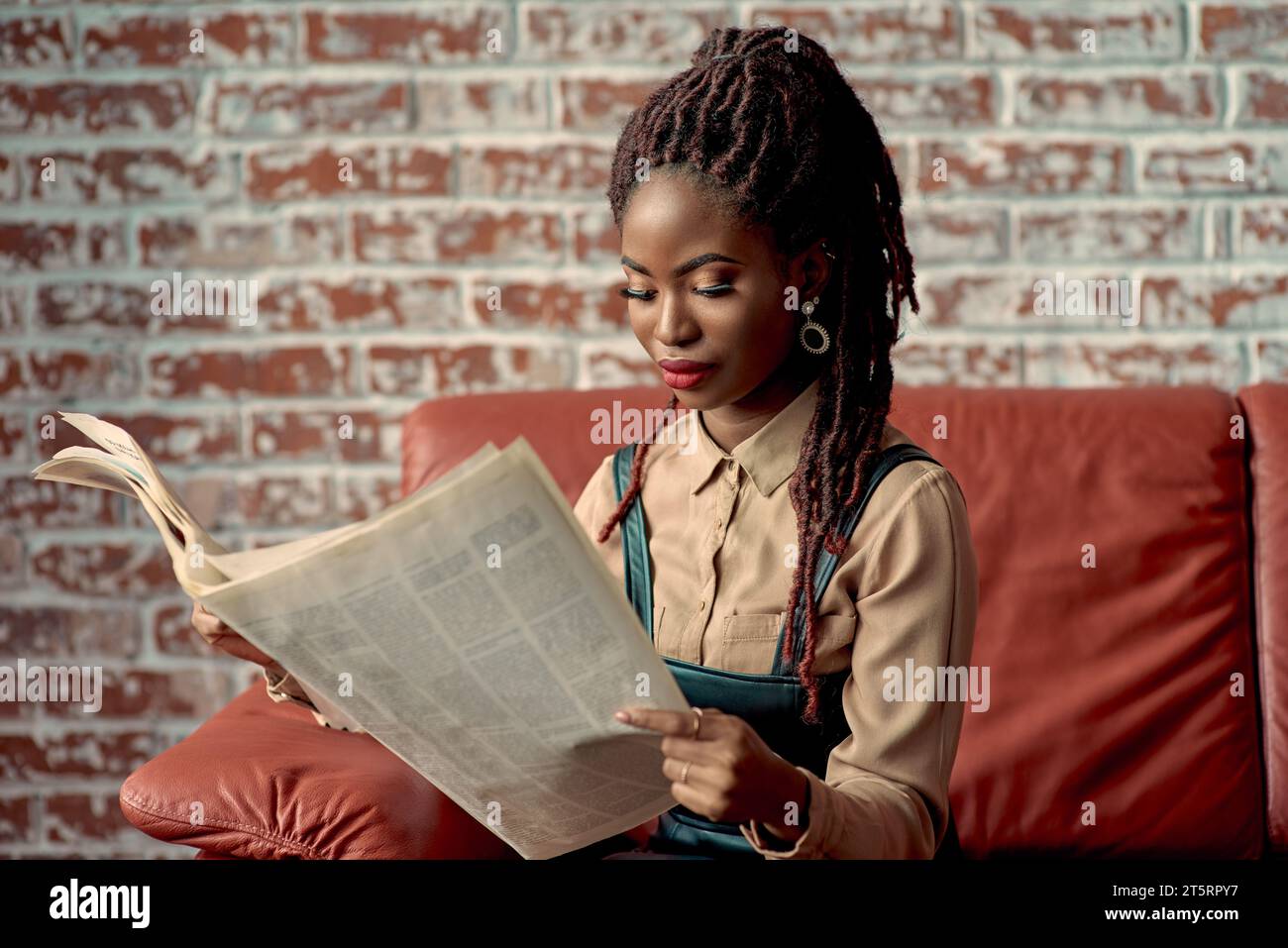Wunderschöne schwarze Frau mit Dreadlocks, die sich darauf konzentrierte, eine Zeitung zu lesen, während sie auf einer roten Ledercouch vor einer roten Ziegelwand mit Kopierraum saß. Y Stockfoto