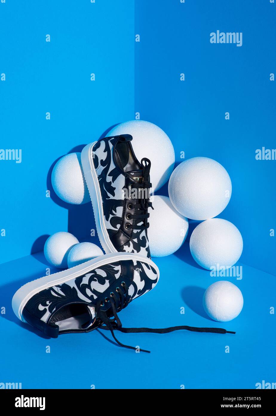 Eine Studioaufnahme mit stylischen lässigen Damen-Sneakers auf blauem Hintergrund in Komposition mit weißen strukturierten Kugeln. Weißes und schwarzes Muster, schwarzer Scho Stockfoto