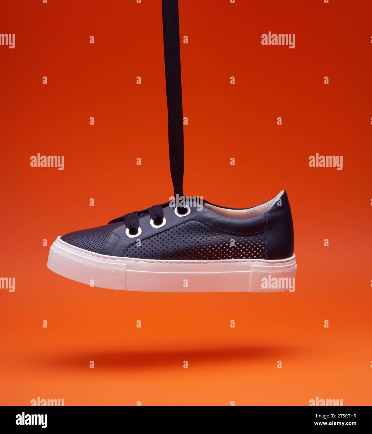 Ein stilvoller, legerer, marineblauer Sneaker, der vor einem leuchtend orangefarbenen Hintergrund an Schnürsenkeln hängt. Perforiertes Leder, dicke weiße Sohle, breite Schnürsenkel, Stockfoto