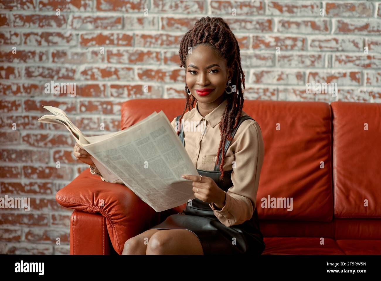 Ziemlich selbstbewusste Afrikanerin, die auf dem roten Sofa sitzt und die Zeitung vor einer Backsteinwand mit Kopierraum liest. Manager beim Mittagessen Stockfoto