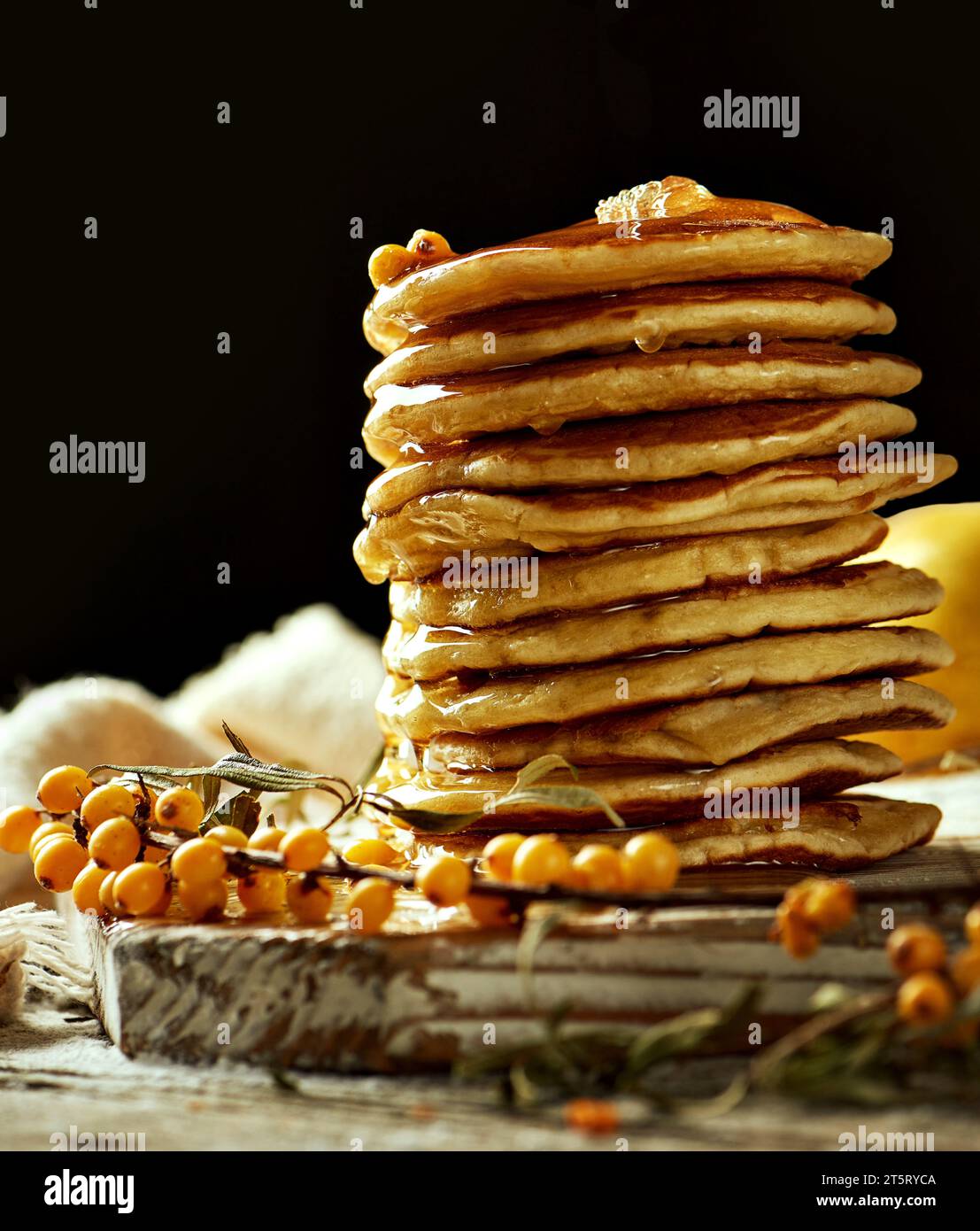 Flauschige Pfannkuchen mit Honig auf einem hellen Holzbrett, mit einem Sanddornzweig und einer Birne auf dem Hintergrund. Kräftige Kontrastfarben. Der Conce Stockfoto
