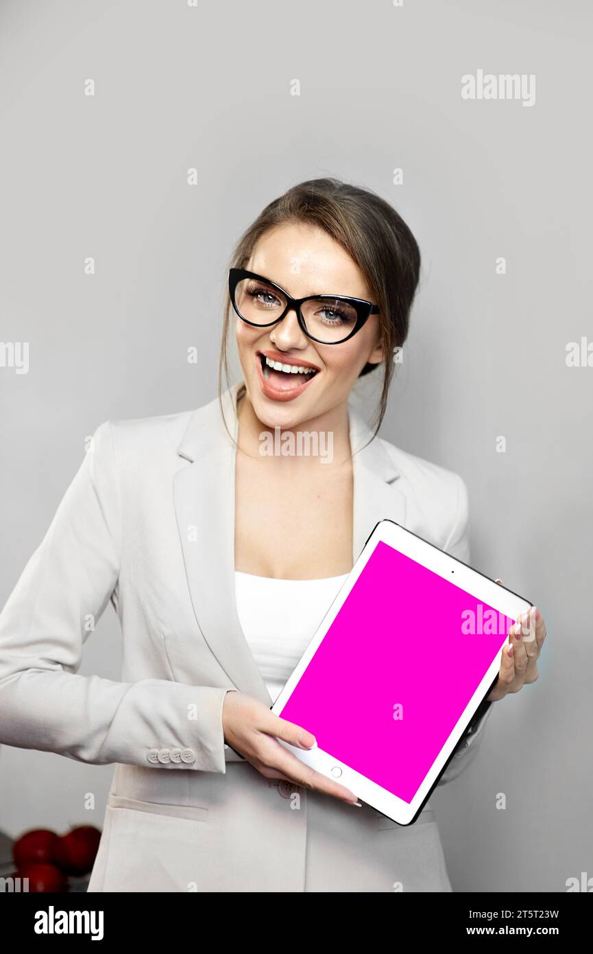 Die schöne junge Frau, die eine Jacke und eine schwarz gerahmte Brille trägt, hat einen aufregenden Gesichtsausdruck, während sie etwas auf einem digitalen Tablet vorführt. Leerer Stift Stockfoto