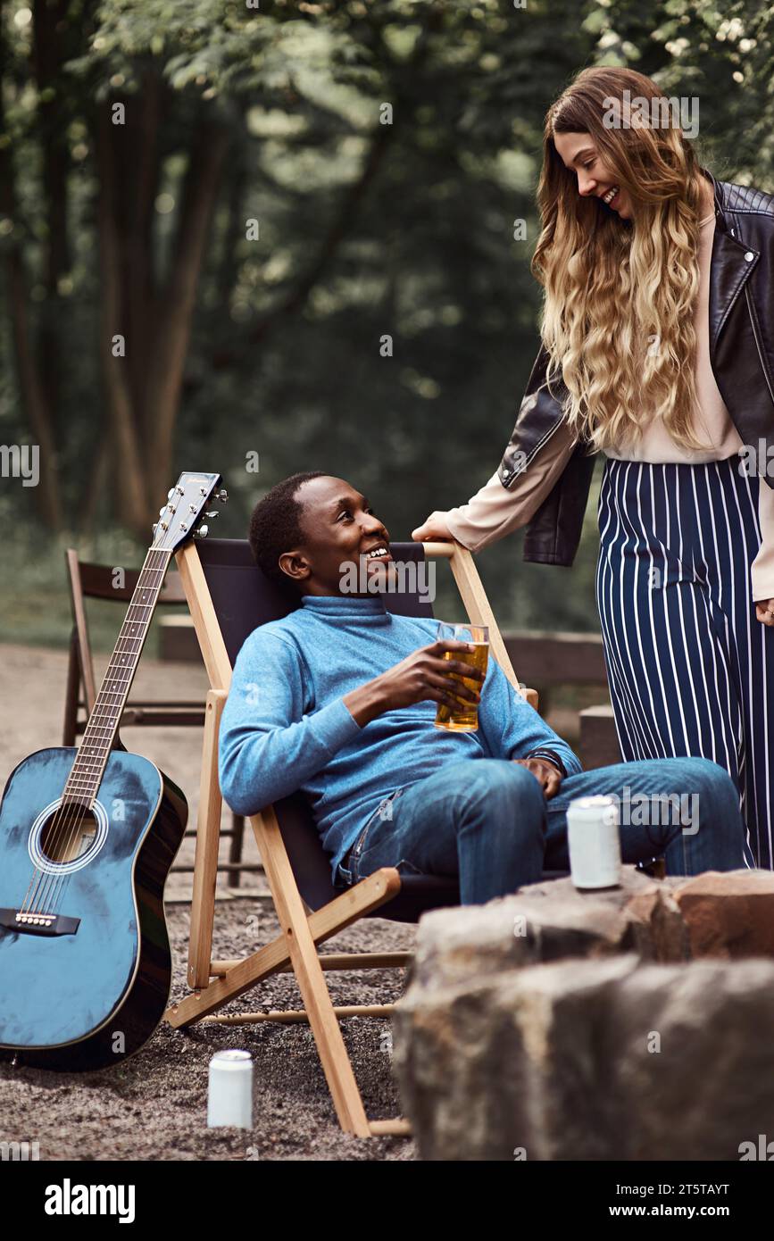 Ein schwarzer Mann sitzt auf einem Campingstuhl und hält ein Glas Bier, während die weiße Frau neben ihm steht und sich gegenseitig anlächelt. Stockfoto