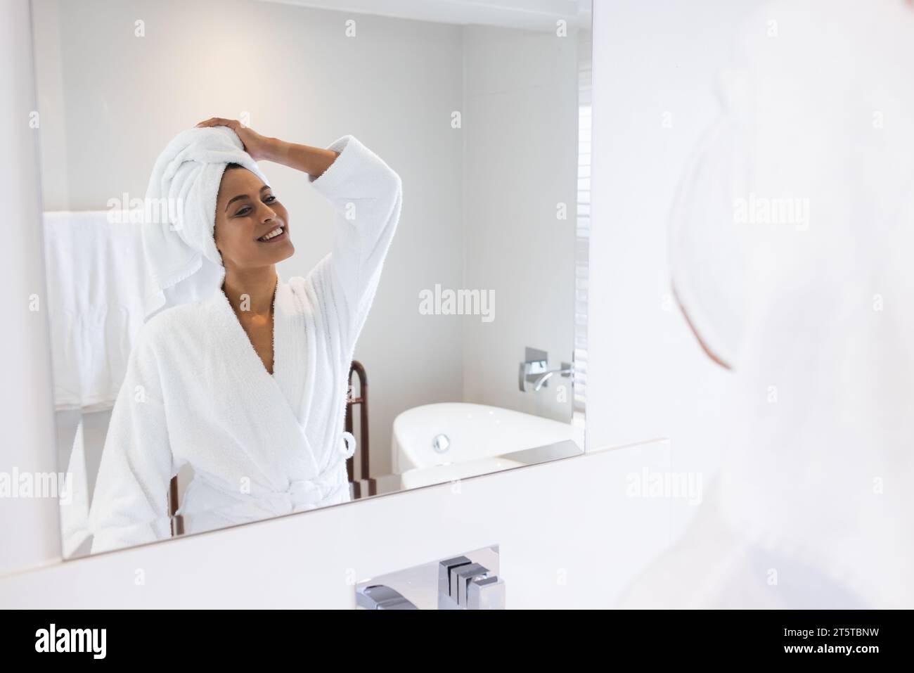 Glückliche Frau mit weißem Bademantel und Handtuch auf dem Kopf vor dem Spiegel im Badezimmer Stockfoto