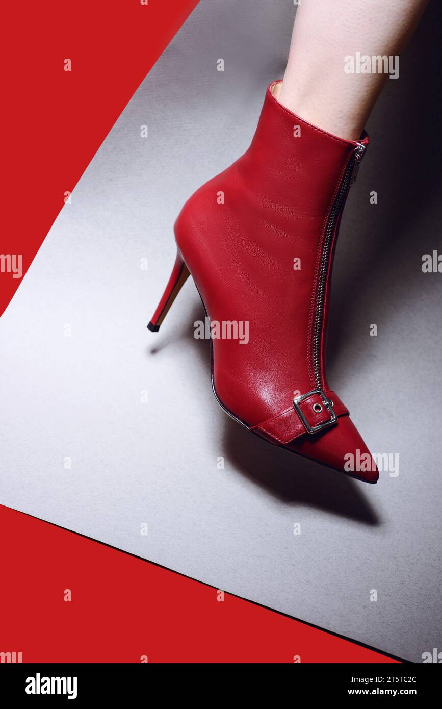 Ein Damenfuß in einem stilvollen dunkelroten Lederstiefel mit hohem Absatz, Reißverschluss und Riemen mit Schnalle vorne auf einem rot-grauen Papierhintergrund. Modell f Stockfoto