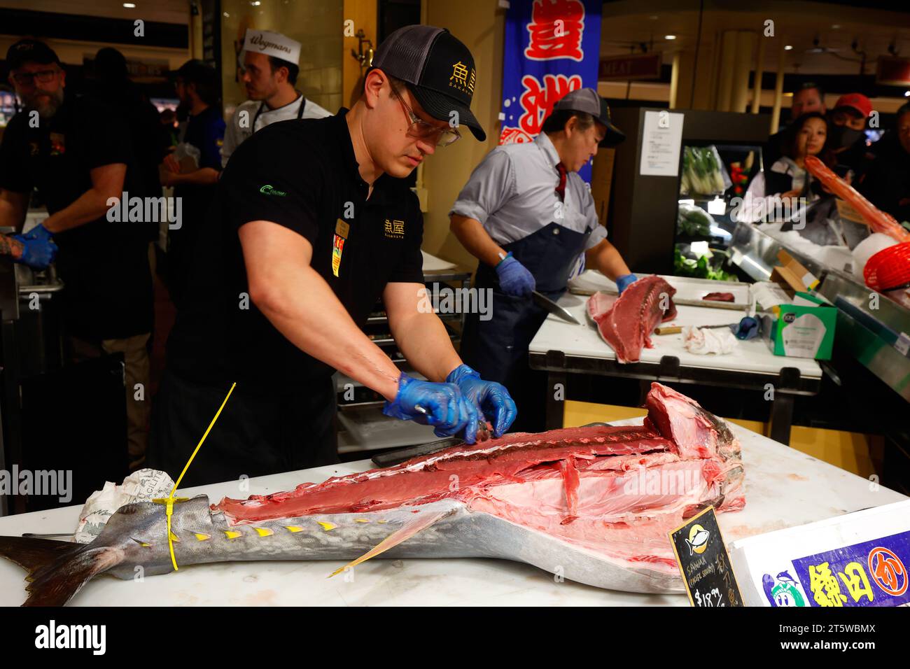 November 2023, New York. Ein Fischhändler kratzt Fleisch von den Rippenknochen (Nakaochi) von einem kleinen wild gefangenen MSC Atlantischen Roten Thun (Thunnus thynnus) Hon maguro; im Hintergrund bereitet ein japanischer Fischhändler Stücke von Akami und Otoro aus einer Thunfischlende zu. Am Sakanaya Fischmarkt im Wegmans Astor Place. An der Fischtheke werden frischer Fisch und Meeresfrüchte aus der Region sowie japanischer Fisch aus dem Toyosu Fish Market in Tokio angeboten. Stockfoto