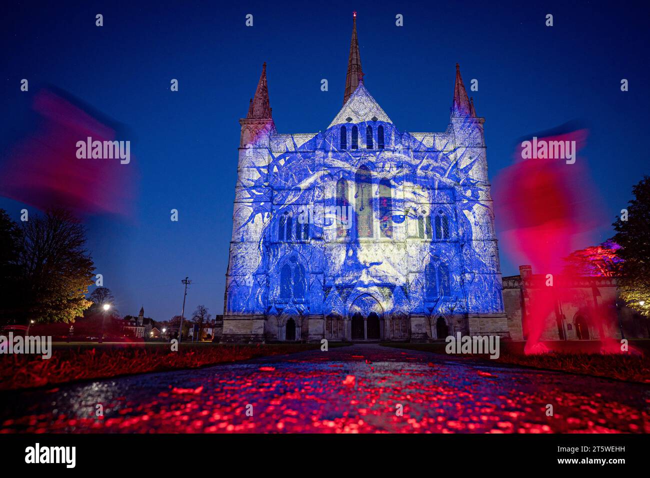 Menschen erzeugen Unschärfen, wenn sie mit langer Verschlusszeit an der Kamera vorbeilaufen, während farbenfrohe Projektionen auf die Salisbury Cathedral in Wiltshire fallen, für die Installation Sarum Lights - Illuminating Art Bilddatum: Montag, 6. November 2023. Stockfoto